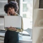 Businesswoman leaning on a windowsill, holding a laptop in a sunlit office.